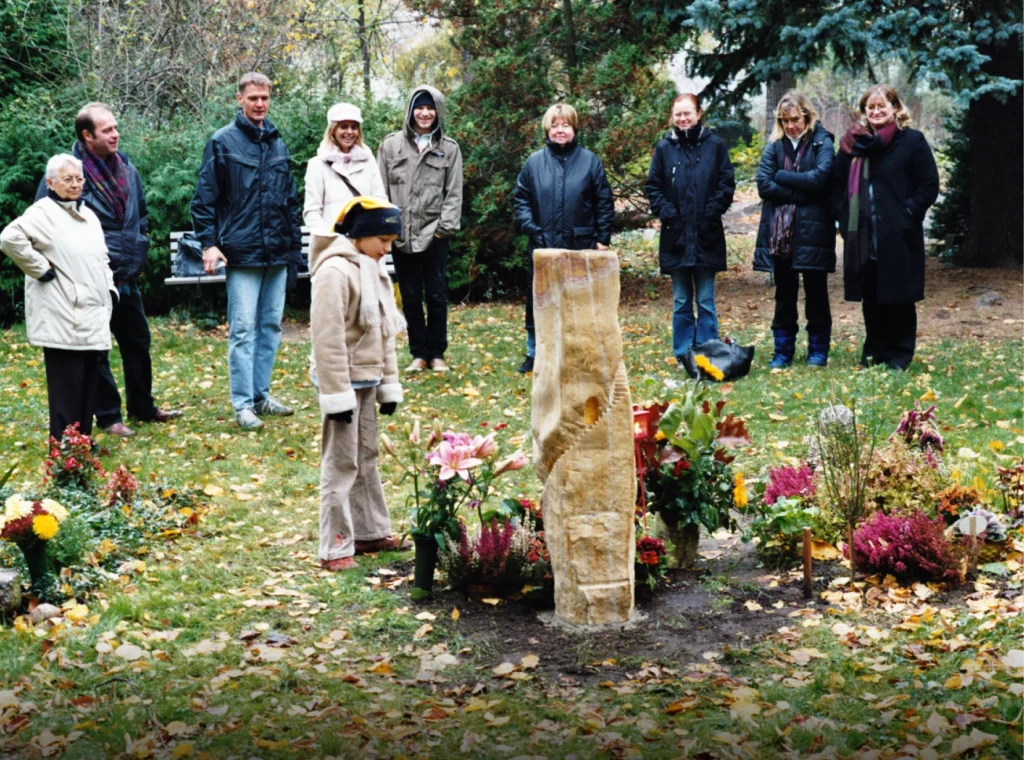 Sandstein-Grabmal mit treppenartiger Form, davor ein Kind und im Hintergrund mehrere Menschen
