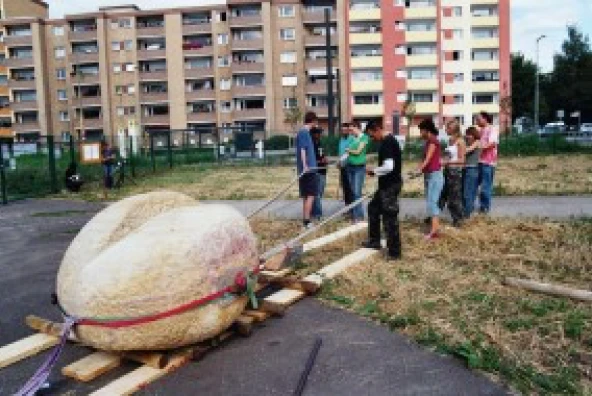 Menschen bewegen eine große Sandstein-Schrippe mit Brechstangen über ein Feld